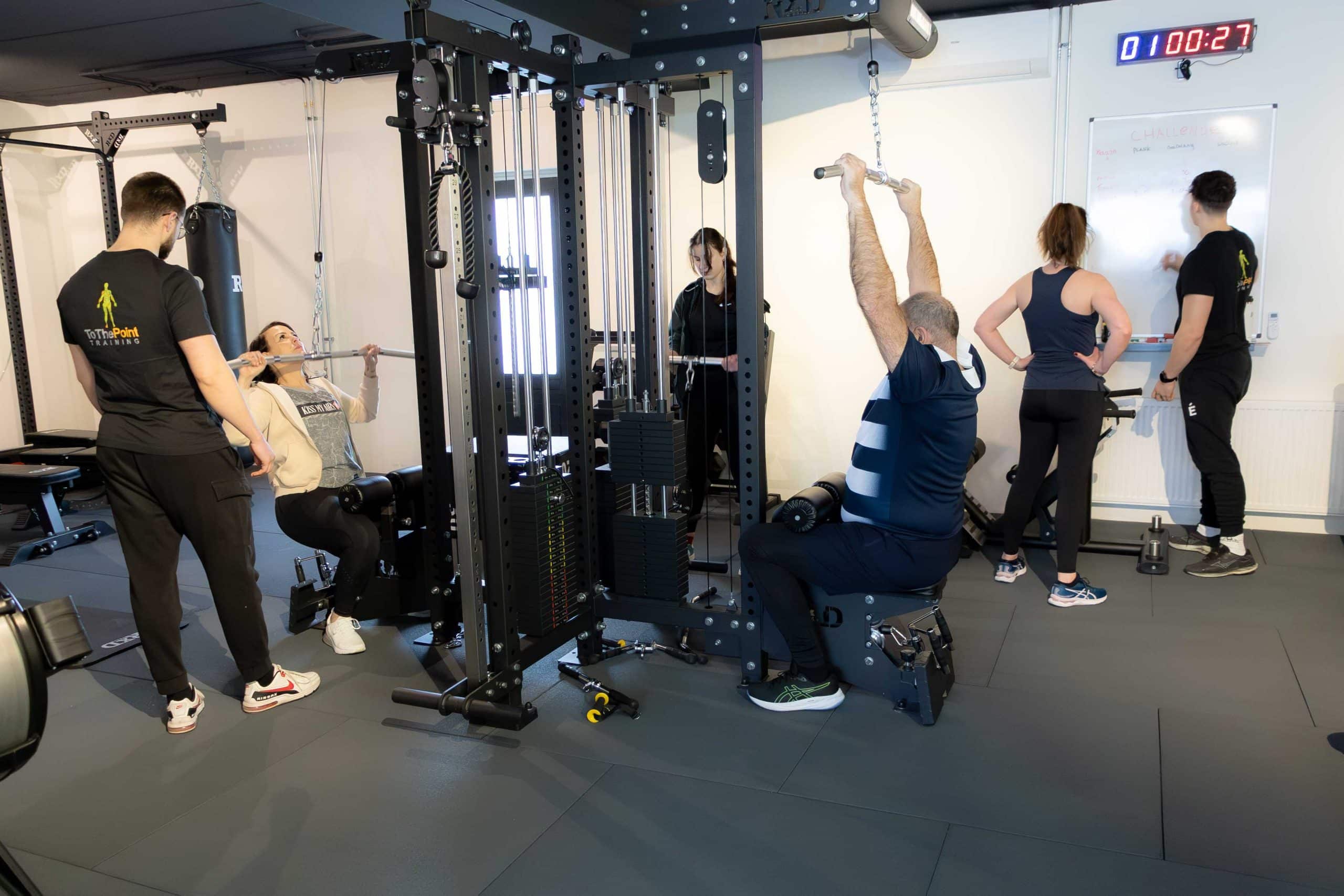 Groepstraining in de gym met personal trainers en deelnemers die krachttrainingsoefeningen uitvoeren bij een kabelstation.